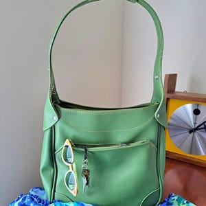 May include: A green shoulder bag with a textured surface, featuring a single top handle, a front zippered pocket, and a set of keys attached to the zipper pull. A pair of yellow-framed sunglasses is tucked into the front pocket of the bag.