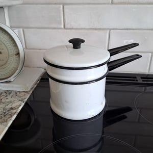 May include: A white enamel double boiler with a black handle and lid knob sits on a black stovetop. The pot has a black trim and is next to a vintage scale with a white face and black numbers.