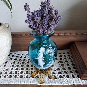 May include: A small, blue glass vase with a white cameo design of a girl holding a ball. The vase has a gold-colored base with three legs and is filled with dried lavender flowers.