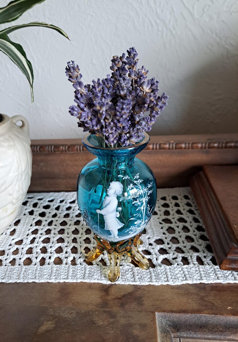 May include: A small, blue glass vase with a white cameo design of a girl holding a ball. The vase has a gold-colored base with three legs and is filled with dried lavender flowers.