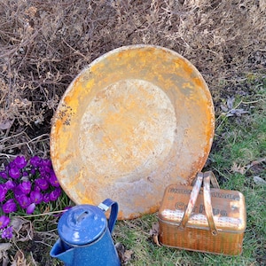 May include: An aged, rusty metal pan and a blue enamelware coffee pot rest on the grass next to a small, vintage picnic basket. Purple crocus flowers bloom nearby. The picnic basket has the words "Union Leader" and "Cup Jug" on it.