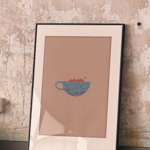 Op de afbeelding: Ingelijste kunstdruk met een minimalistische illustratie van een blauwe kop met twee rode bollen erin. Het kunstwerk staat tegen een gedempte perzikachtergrond, is zwart omlijst en leunt tegen een getextureerde muur.