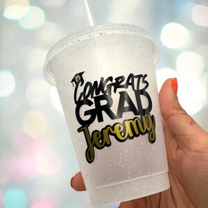 May include: A white glitter tumbler with a black and gold sticker that says "Congrats Grad Jeremy".