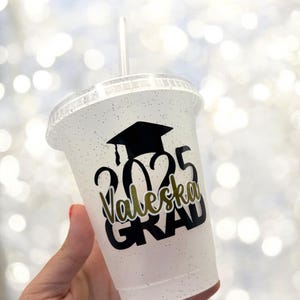 May include: A white plastic cup with a black glitter finish. The cup has a black graduation cap, the year 2025, the name "Valeska" and the word "GRAD" printed on it.