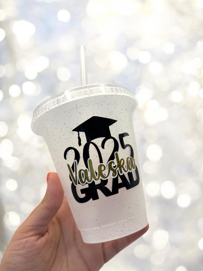 May include: A white plastic cup with a black glitter finish. The cup has a black graduation cap, the year 2025, the name "Valeska" and the word "GRAD" printed on it.