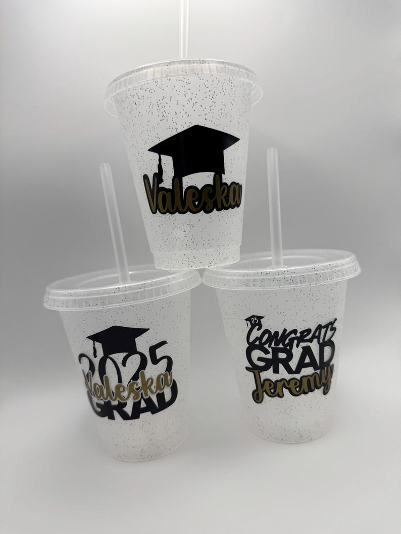 May include: Three clear plastic cups with glitter, each with a different graduation design. One cup has a black graduation cap and the name "Valeska" in gold lettering. Another cup has the year "2025" and the name "Valeska" in gold lettering. The third cup has the text "Congrats Grad Jeremy" in black lettering.