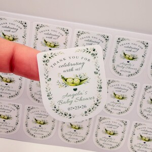 A hand peels a customized sticker featuring an illustrated pea pod from a sheet of printed baby shower labels.