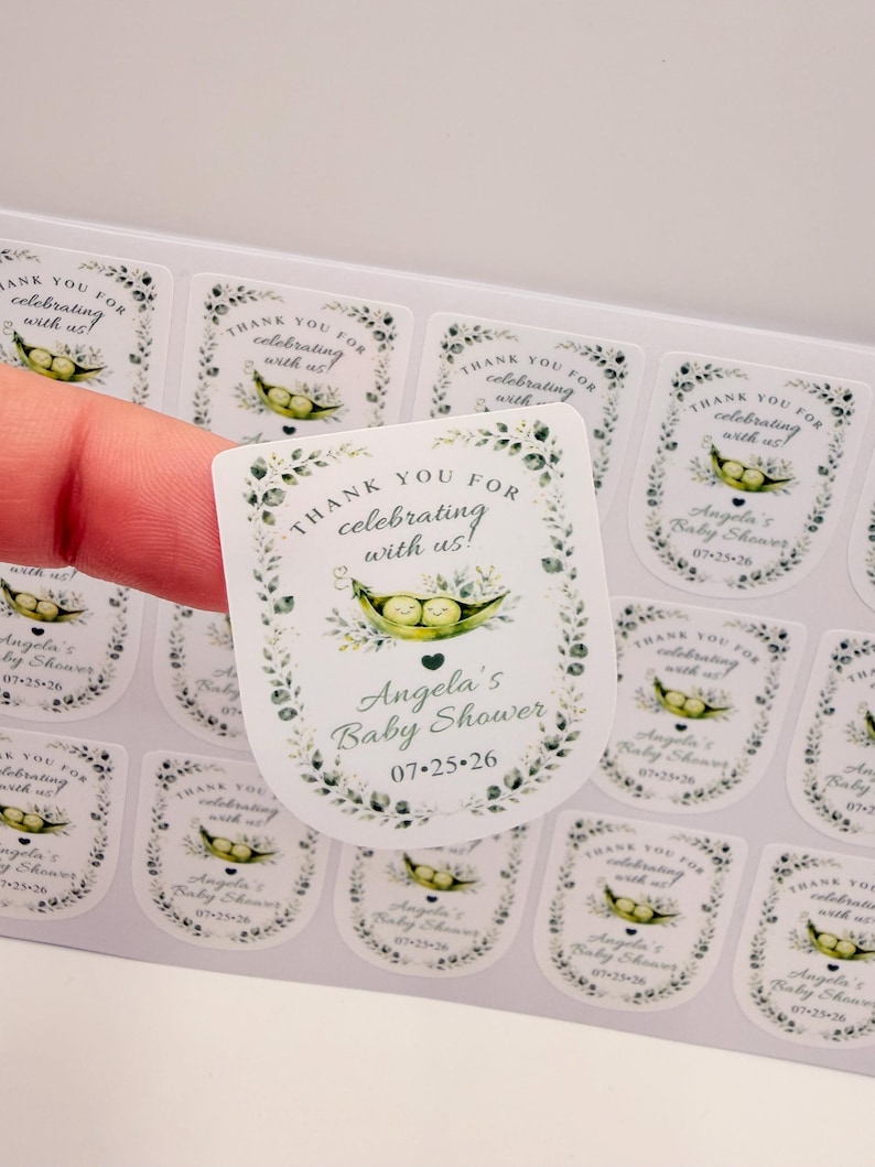 A hand peels a customized sticker featuring an illustrated pea pod from a sheet of printed baby shower labels.