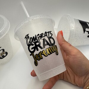 May include: A white glitter tumbler with a black and gold design that says "Congrats Grad Jeremy".
