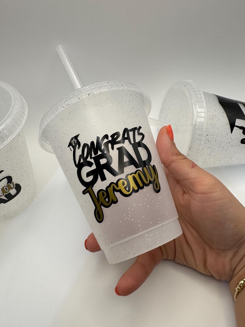 May include: A white glitter tumbler with a black and gold design that says "Congrats Grad Jeremy".