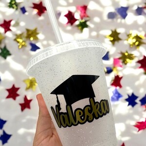 May include: A white glitter tumbler with a black graduation cap and gold lettering that says "Valeska".