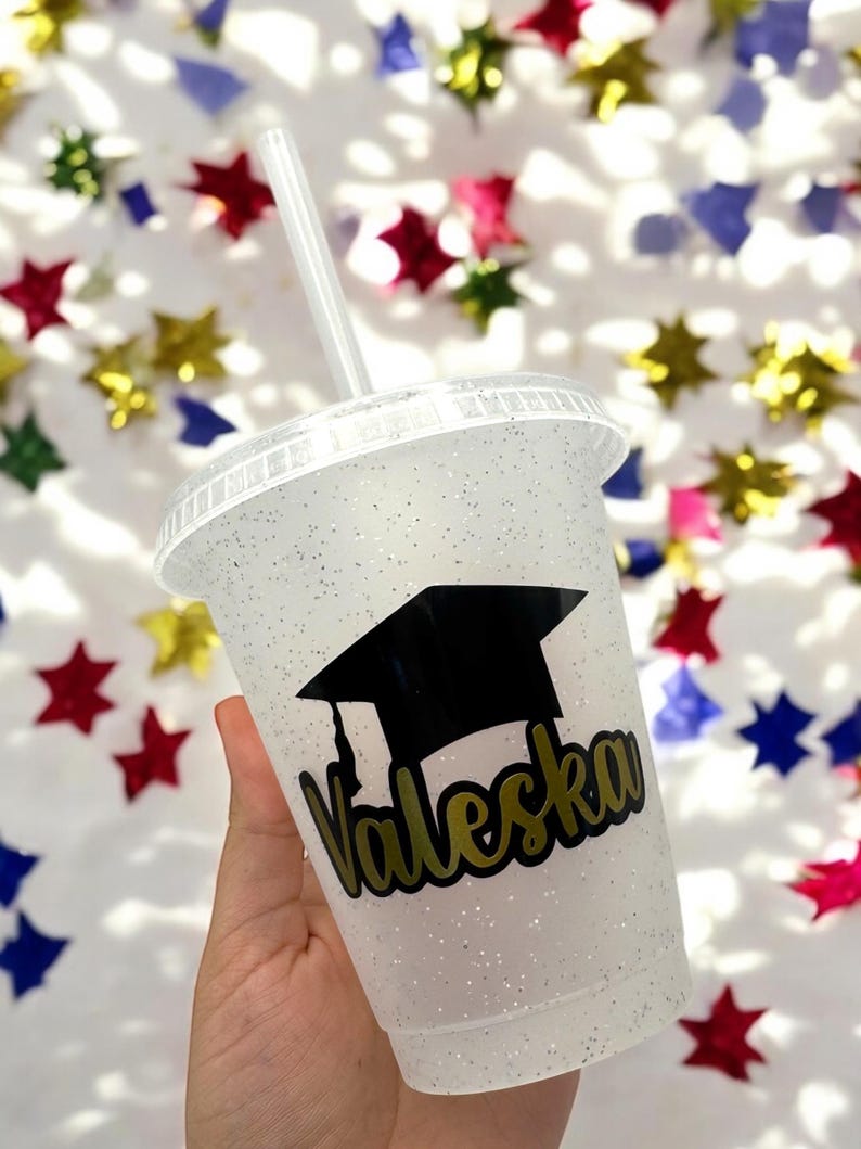 May include: A white glitter tumbler with a black graduation cap and gold lettering that says "Valeska".
