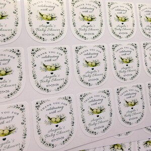 A close-up overhead view displays a sheet of glossy sticker labels printed with a pea in a pod baby shower design.