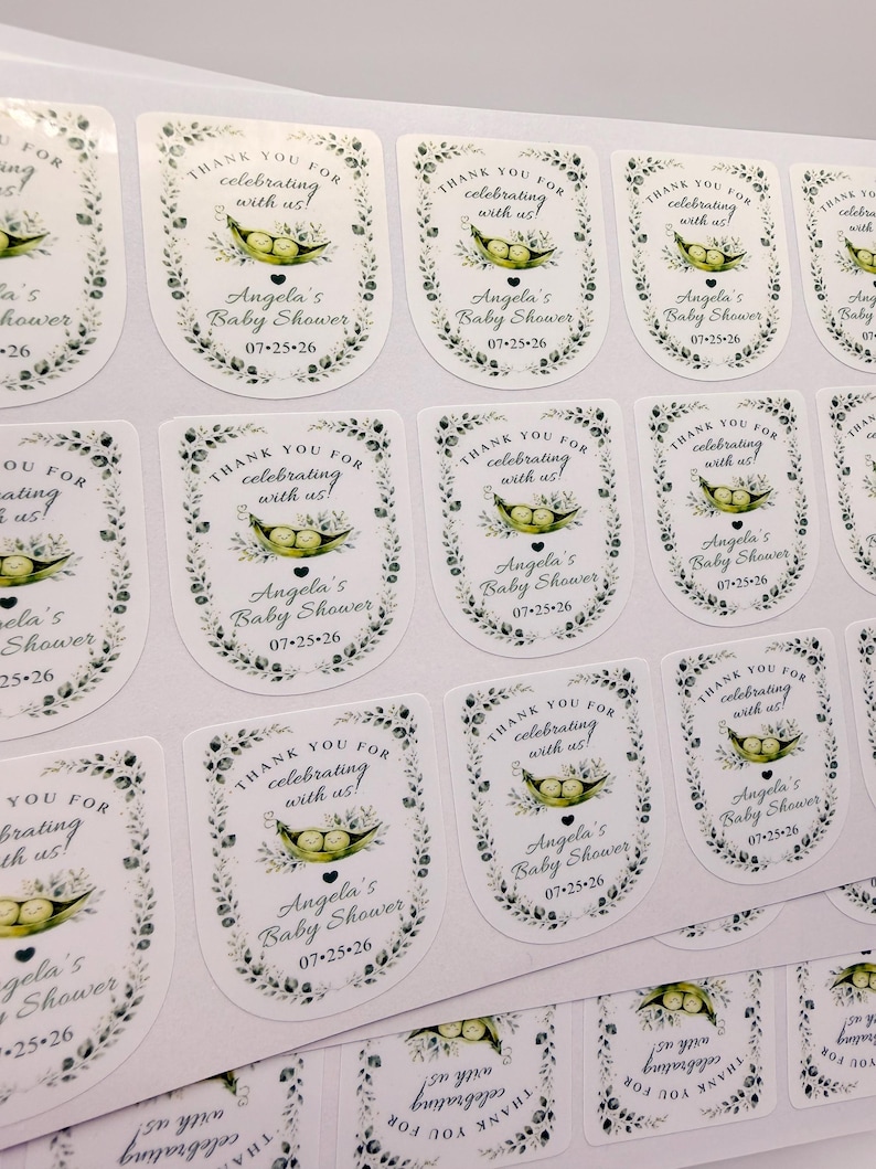 A close-up overhead view displays a sheet of glossy sticker labels printed with a pea in a pod baby shower design.