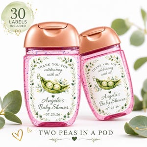 Two hand sanitizer bottles containing pink glitter and topped with rose gold caps feature custom pea pod baby shower labels against a white and green backdrop.
