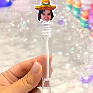 May include: A clear plastic bubble wand with a smiling child's face wearing a sombrero. The wand is held in a hand, and the background is filled with bubbles. The sombrero is yellow, red, and green.