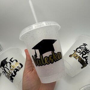 May include: Three clear plastic cups with glitter and black graduation caps. The cups have gold lettering with the names "Valeska", "Jeremy", and "Valeska Grad 2023".