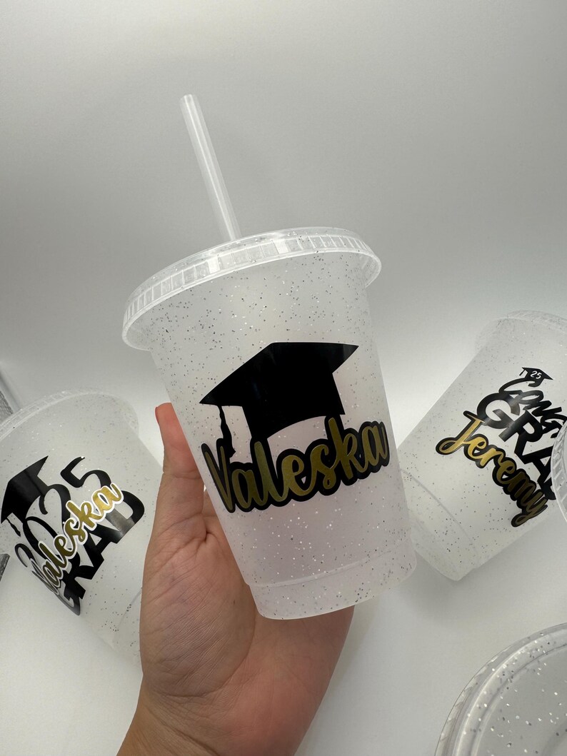 May include: Three clear plastic cups with glitter and black graduation caps. The cups have gold lettering with the names "Valeska", "Jeremy", and "Valeska Grad 2023".