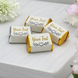 Puede incluir: Cinco chocolates envueltos individualmente con extremos de papel dorado y etiquetas blancas. Cada etiqueta tiene el texto "Your Text" en dorado, junto con un diseño de corazón y hojas. Los chocolates están dispuestos sobre una superficie blanca, con flores en el fondo.