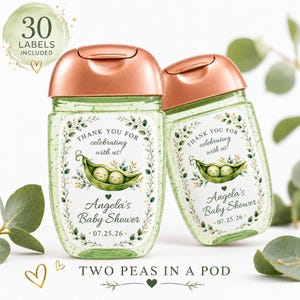 Two clear hand sanitizer bottles with rose gold caps and green glitter gel, decorated with custom pea in a pod labels, are displayed against a white background with eucalyptus leaves.