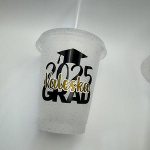 May include: White glitter tumbler with a black graduation cap, the year 2025, the name "Valeska" and the word "GRAD" in gold lettering.