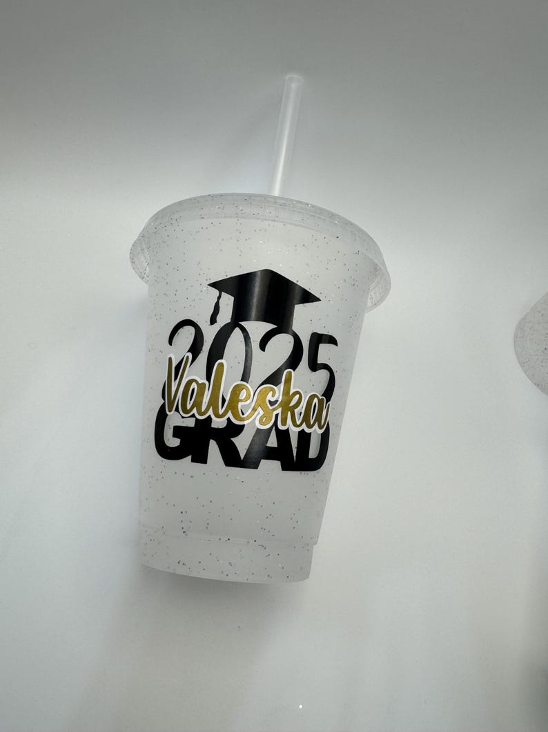 May include: White glitter tumbler with a black graduation cap, the year 2025, the name "Valeska" and the word "GRAD" in gold lettering.