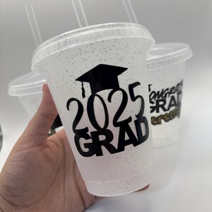 May include: Clear plastic cups with glitter and black vinyl lettering that says "2023 Grad" and a graduation cap. The cups have black straws.