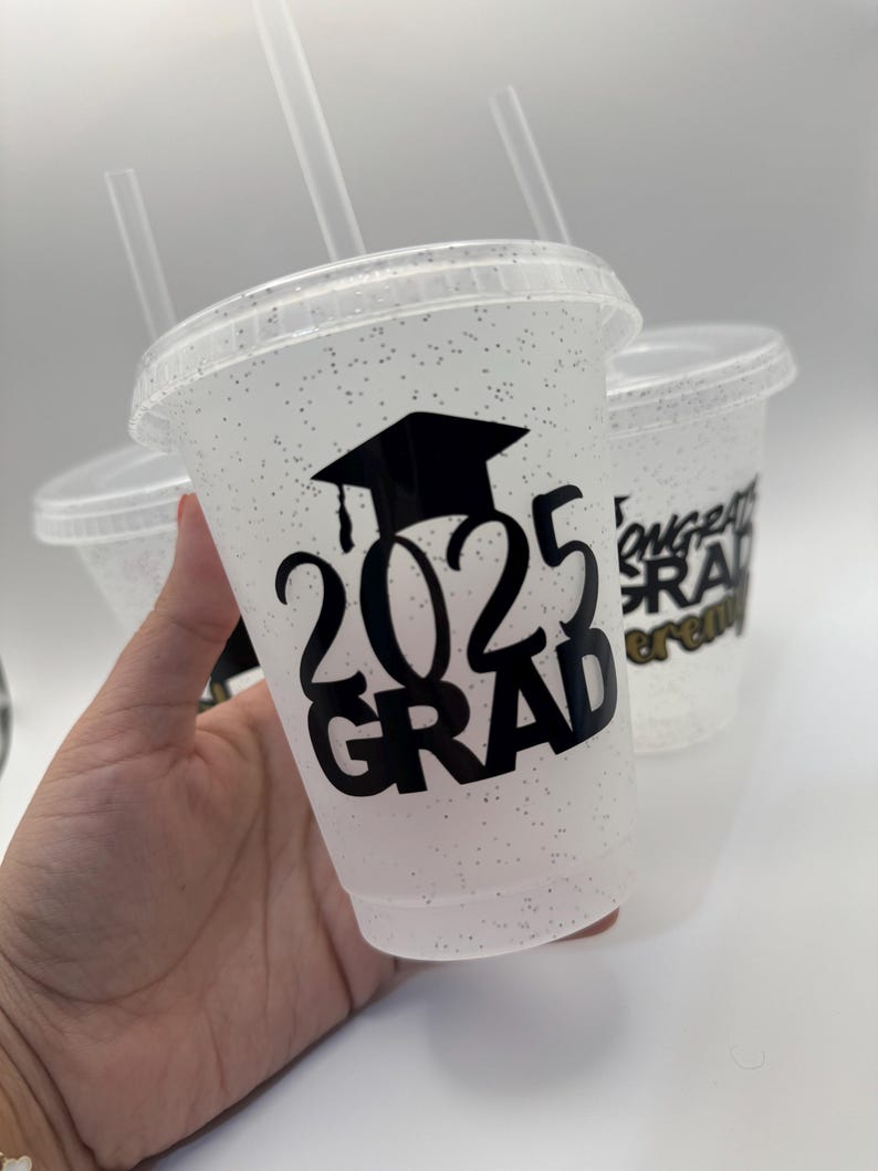 May include: Clear plastic cups with glitter and black vinyl lettering that says "2023 Grad" and a graduation cap. The cups have black straws.