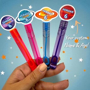 May include: Four colorful bubble wands in red, pink, blue, and purple, each with a matching cap and bubble loop. The wands have stickers that say "ARMAAN TURNS 6" and the text "Your custom Name & Age!" on a blue background with stars.