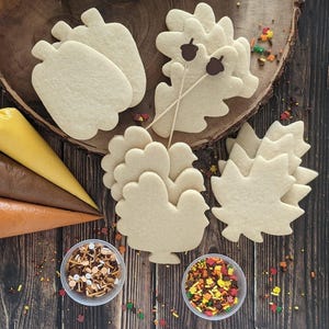 May include: An assortment of unadorned sugar cookies in the shapes of pumpkins, oak leaves, turkeys, and maple leaves. Two small bowls of sprinkles and three piping bags filled with yellow, brown, and orange icing are also present.