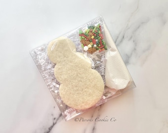 May include: A snowman-shaped cookie kit in a clear plastic box. The kit includes a large, undecorated cookie, a bag of colorful sprinkles, and a bag of white icing. The text "Parsons Cookie Co" is printed on the bottom.