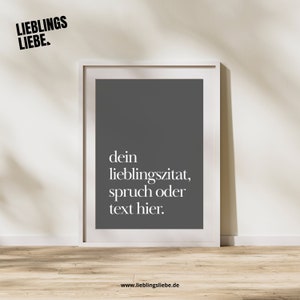 May include: A framed print with a dark gray background and white text that reads "dein Lieblingszitat, spruch oder text hier." The top left corner has the text "LIEBLINGS LIEBE." in black. The frame is white, and the background is a light beige wall.
