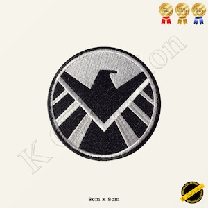 Puede incluir: Parche bordado en blanco y negro del logotipo de S.H.I.E.L.D. El logotipo es un círculo con un águila blanca en el interior y una estrella blanca en el centro. El círculo está rodeado por un anillo blanco con un borde negro. El parche mide 8 cm x 8 cm.