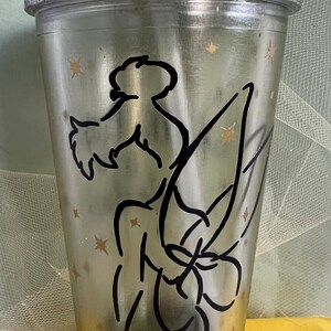 Peut inclure: Un gobelet en plastique transparent avec un bord et une base en paillettes argent&eacute;es. Le gobelet pr&eacute;sente une silhouette noire de f&eacute;e avec des ailes et un motif d'&eacute;toiles sur le c&ocirc;t&eacute;.