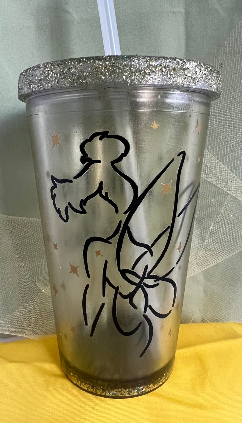 Peut inclure: Un gobelet en plastique transparent avec un bord et une base en paillettes argent&eacute;es. Le gobelet pr&eacute;sente une silhouette noire de f&eacute;e avec des ailes et un motif d'&eacute;toiles sur le c&ocirc;t&eacute;.