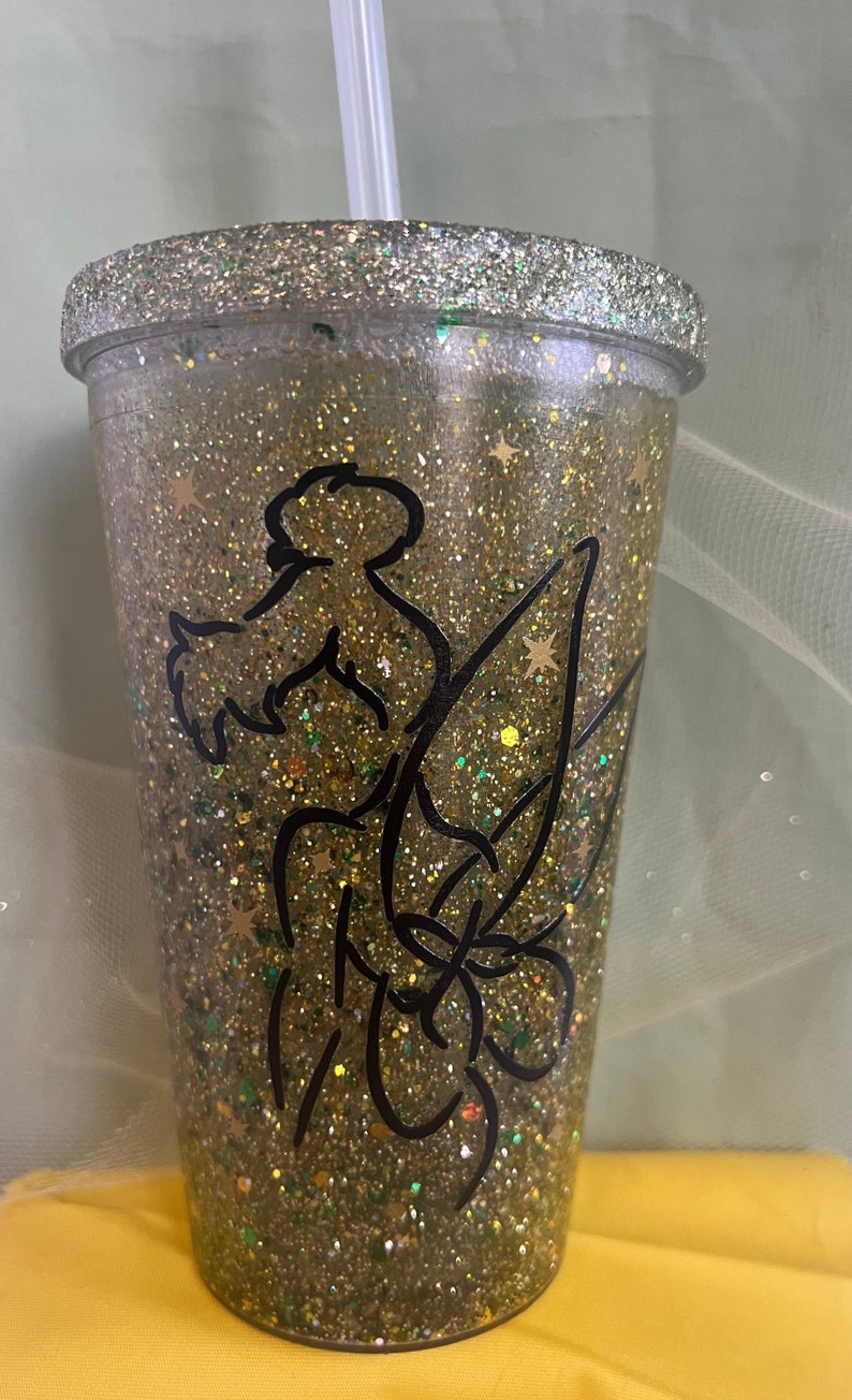 Peut inclure: Un gobelet en paillettes dor&eacute;es avec une silhouette noire de f&eacute;e avec des ailes et des &eacute;toiles. Le gobelet a un couvercle en plastique transparent et une paille en plastique transparente.