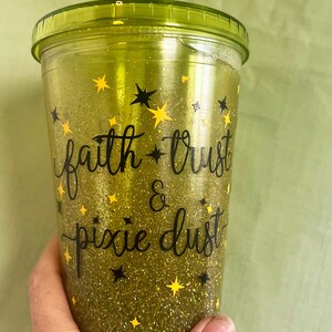 Peut inclure: Un gobelet r&eacute;utilisable vert avec une base en paillettes dor&eacute;es et un lettrage noir qui dit "faith * trust & pixie dust". Le gobelet a une paille jaune et est d&eacute;cor&eacute; d'&eacute;toiles noires et jaunes.