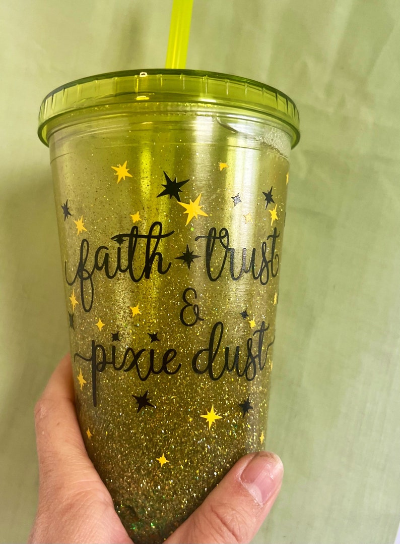 Peut inclure: Un gobelet r&eacute;utilisable vert avec une base en paillettes dor&eacute;es et un lettrage noir qui dit "faith * trust & pixie dust". Le gobelet a une paille jaune et est d&eacute;cor&eacute; d'&eacute;toiles noires et jaunes.