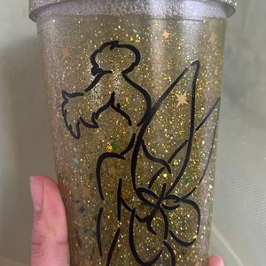Peut inclure: Un gobelet en plastique transparent avec un motif de paillettes dor&eacute;es et une silhouette noire de la f&eacute;e Clochette. Le gobelet est dot&eacute; d'une paille et est parfait pour contenir des boissons.