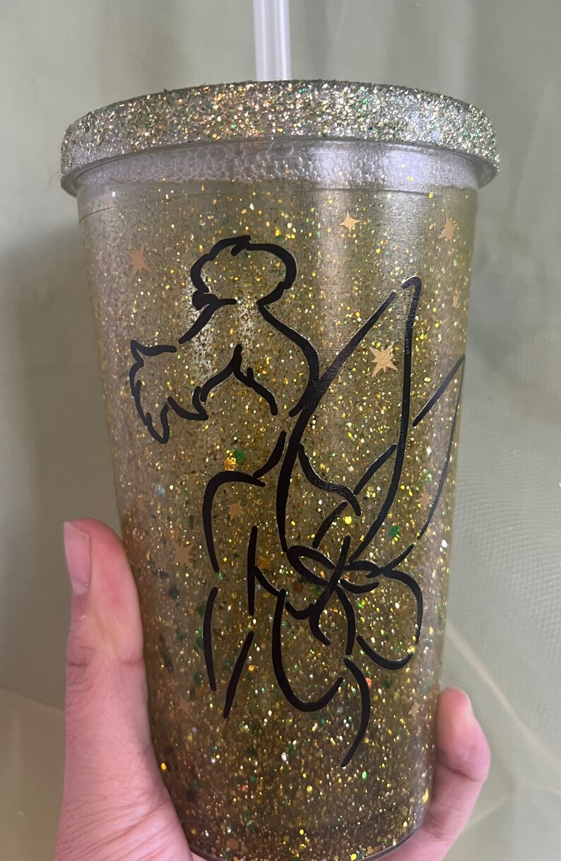 Peut inclure: Un gobelet en plastique transparent avec un motif de paillettes dor&eacute;es et une silhouette noire de la f&eacute;e Clochette. Le gobelet est dot&eacute; d'une paille et est parfait pour contenir des boissons.