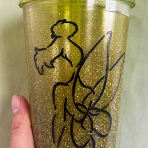 Peut inclure: Un gobelet en paillettes dor&eacute;es avec une silhouette noire de f&eacute;e avec des ailes et une baguette magique. Le gobelet a un couvercle vert et une paille transparente.