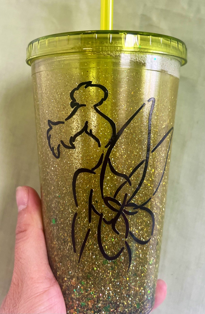 Peut inclure: Un gobelet en paillettes dor&eacute;es avec une silhouette noire de f&eacute;e avec des ailes et une baguette magique. Le gobelet a un couvercle vert et une paille transparente.