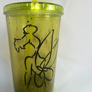 Peut inclure: Un gobelet vert &agrave; paillettes avec un contour noir de la f&eacute;e Clochette. Le gobelet a un couvercle transparent et une paille jaune.