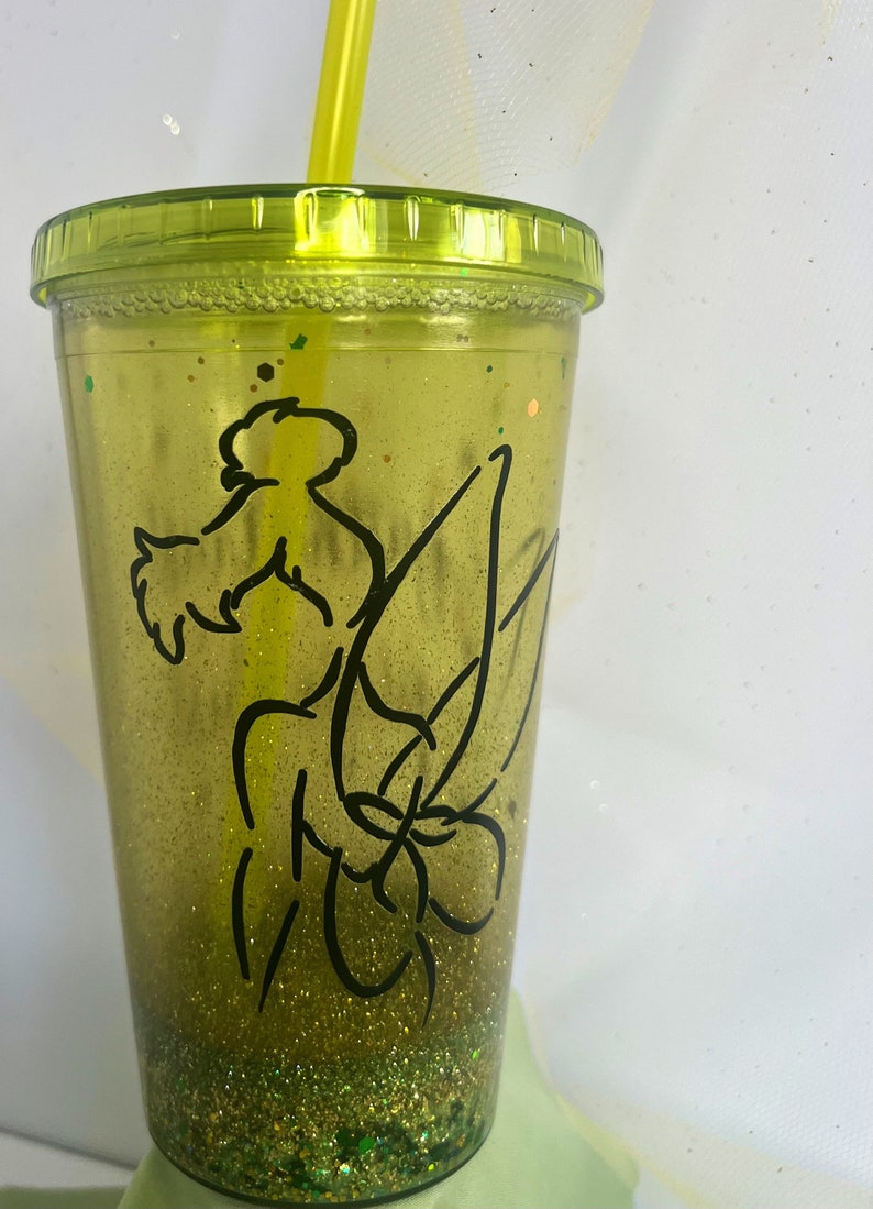 Peut inclure: Un gobelet vert &agrave; paillettes avec un contour noir de la f&eacute;e Clochette. Le gobelet a un couvercle transparent et une paille jaune.