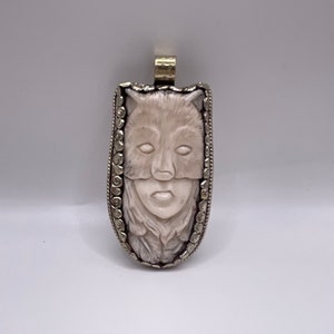 May include: A silver pendant with a carved white wolf face, framed by a silver border with a decorative pattern.