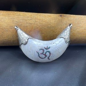 Pode incluir: Um pingente branco em forma de crescente com detalhes prateados e um símbolo Om no centro. O símbolo Om é decorado com pedras turquesa e vermelhas.