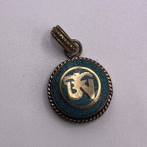 May include: A gold pendant with a turquoise blue inlay featuring a stylized symbol. The pendant is round and has a rope-like border.