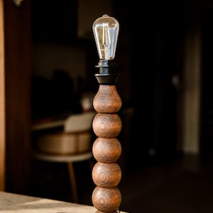 May include: A table lamp featuring a vertical stack of six dark brown wooden spheres on a circular base. The lamp is topped with a black socket and a clear glass Edison-style light bulb with visible internal filaments, creating a tall, slender profile.
