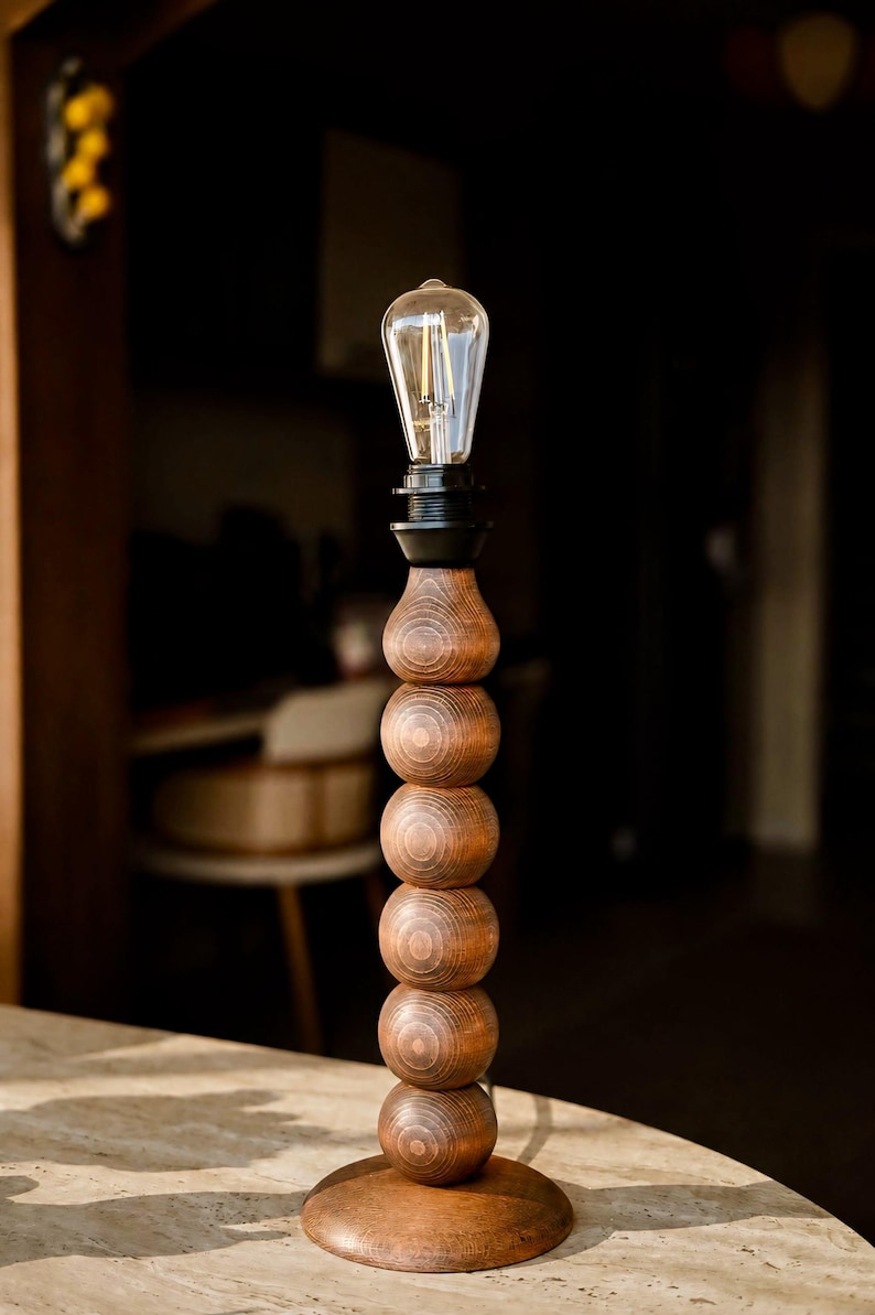 May include: A table lamp featuring a vertical stack of six dark brown wooden spheres on a circular base. The lamp is topped with a black socket and a clear glass Edison-style light bulb with visible internal filaments, creating a tall, slender profile.