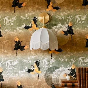 May include: A wall-mounted light fixture with a white lampshade and fringe is set against a wallpaper featuring a repeating pattern of black and gold cranes and bamboo leaves. A small wooden table holds books and a bird figurine.